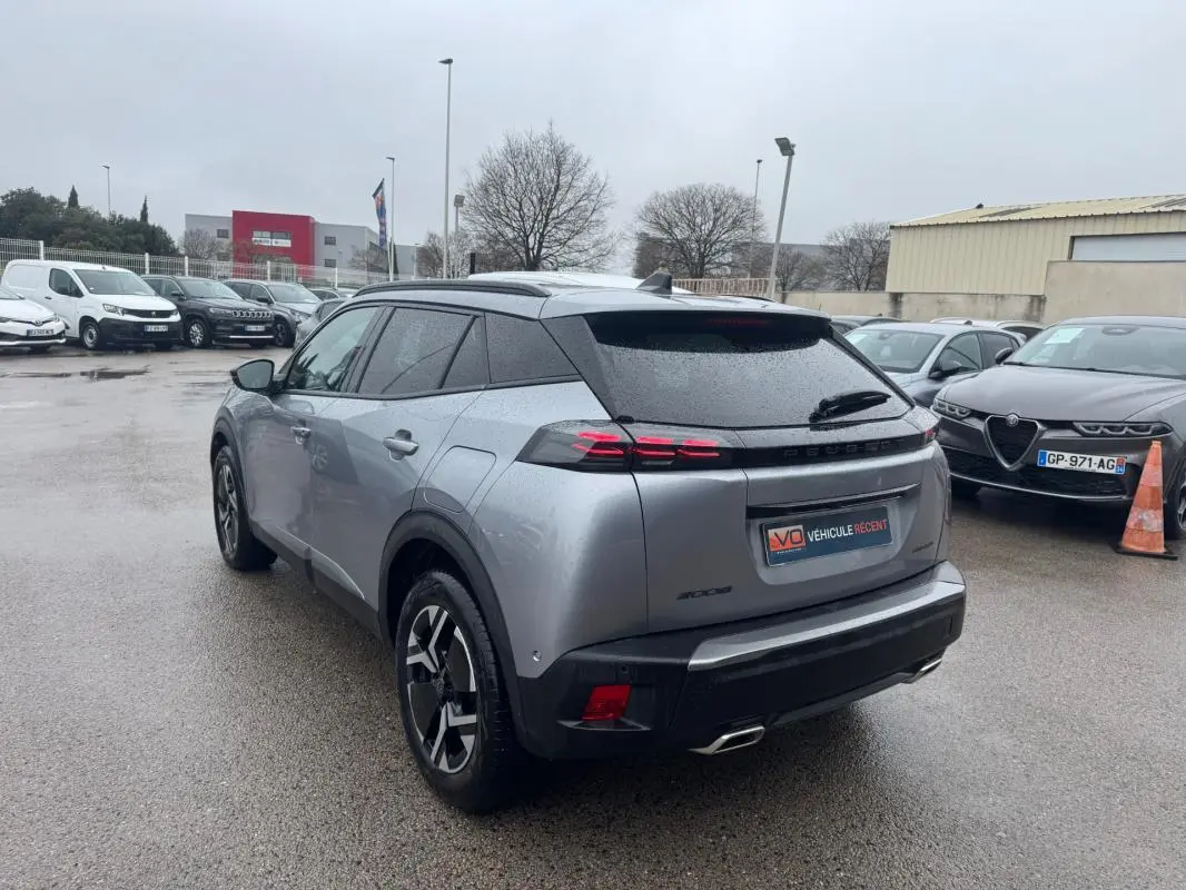 Vue 3/4 arrière gauche d’un Peugeot 2008 Hybrid gris avec jantes alliage 17 pouces et barres de toit visibles par temps humide.