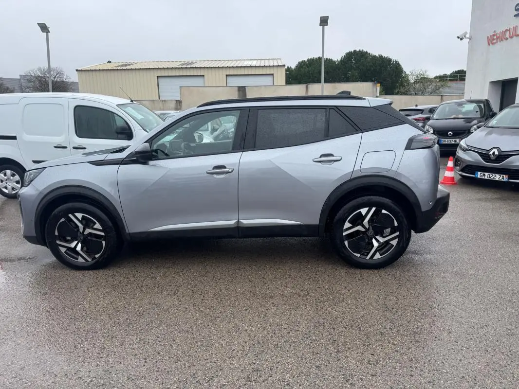 Profil côté gauche d’un Peugeot 2008 Hybrid 145 gris avec barres de toit et jantes alliage 17 pouces, sur parking humide.