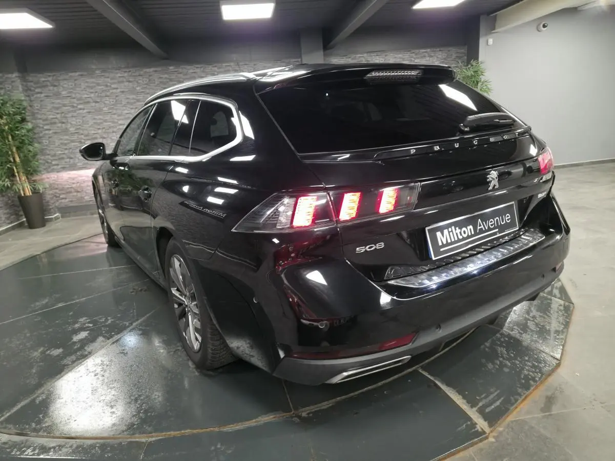 Peugeot 508 SW noire vue 3/4 arrière droit, feux arrière allumés et barres de toit visibles.