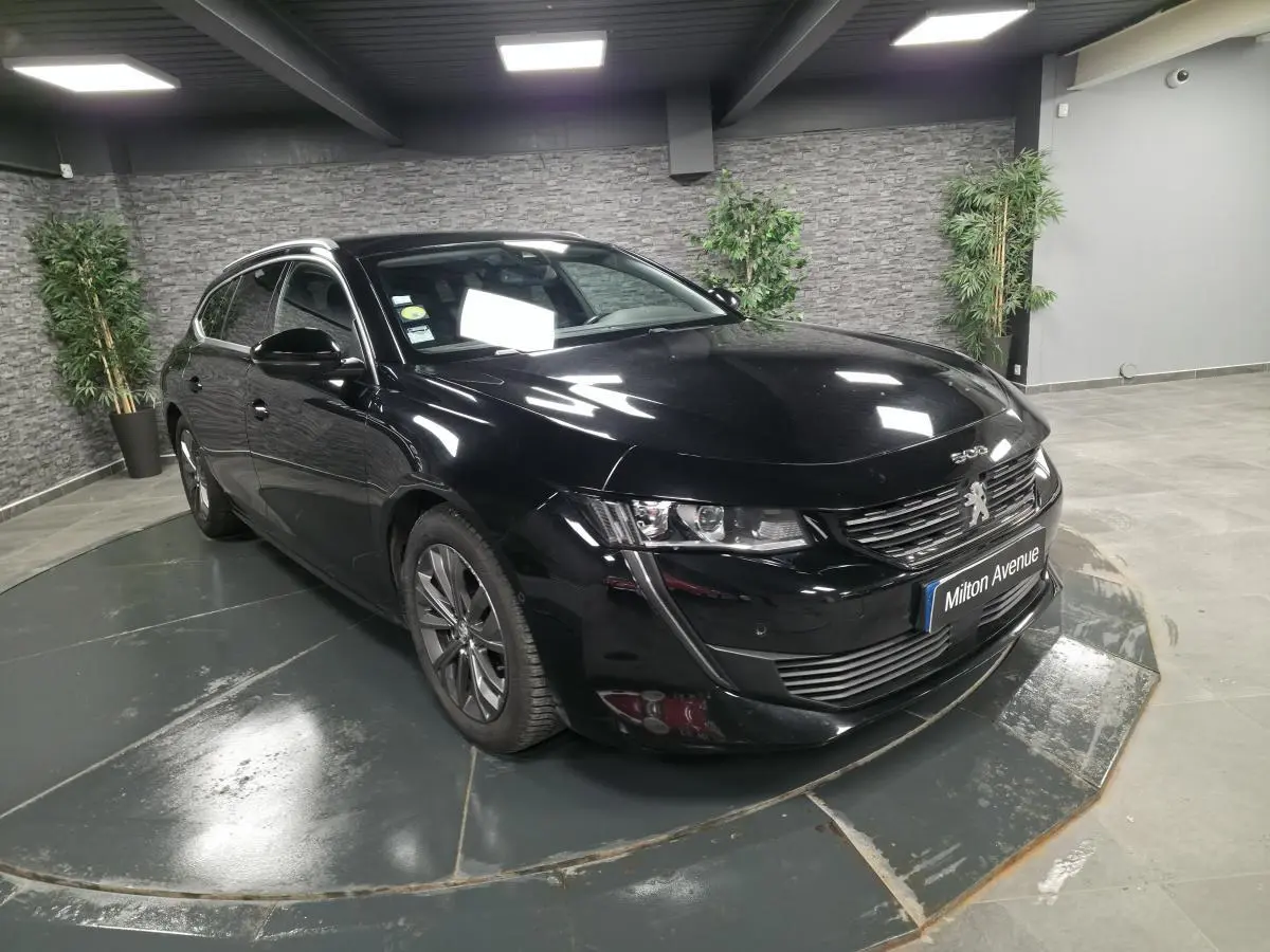 Peugeot 508 SW noire vue 3/4 avant droit avec calandre chromée et barres de toit aluminium en intérieur showroom.