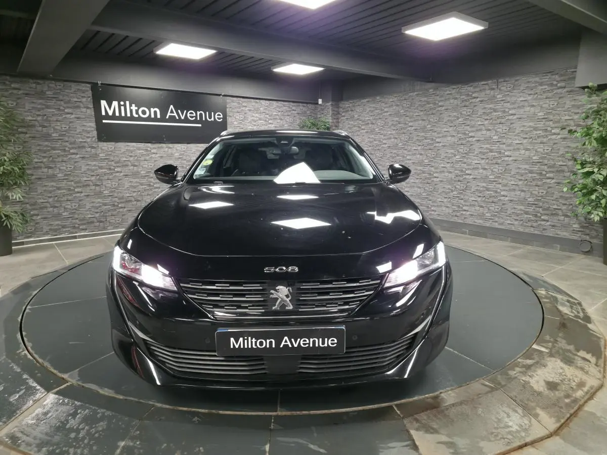 Peugeot 508 SW noire vue de face dans un showroom, avec calandre chromée et phares allumés.