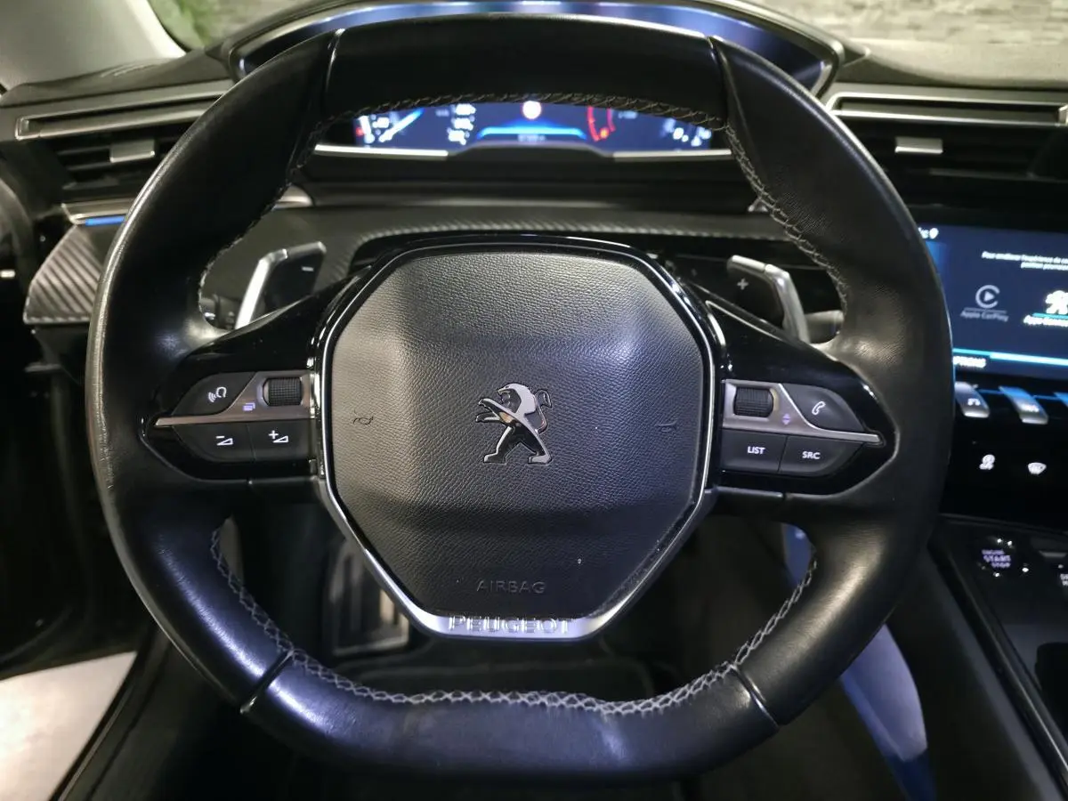 Vue rapprochée du volant cuir noir du Peugeot 508 SW 2019, avec commandes intégrées et tableau de bord numérique en arrière-plan.
