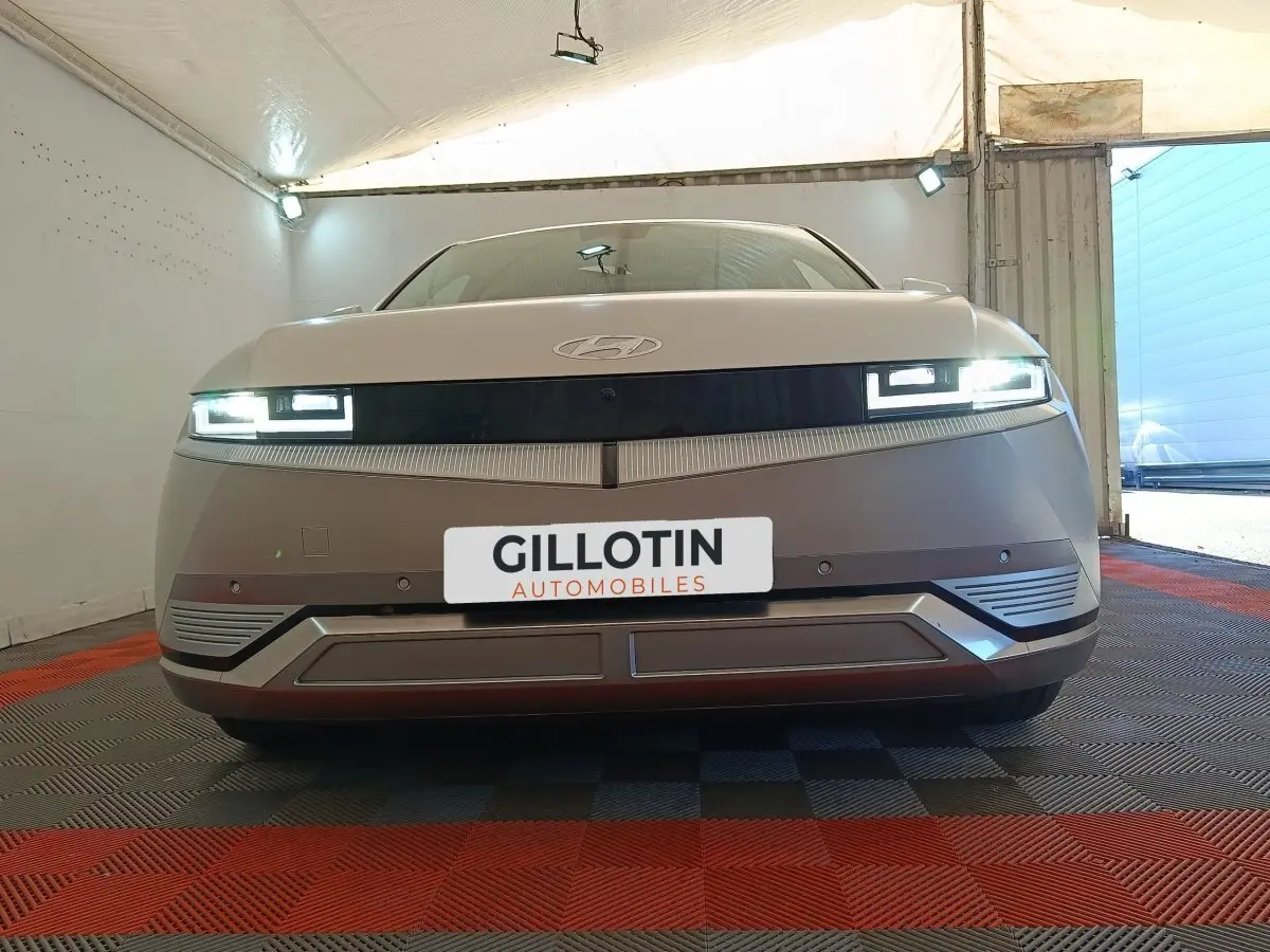 Vue frontale d'une Hyundai IONIQ BEV 77KWH AWD Executive beige avec phares LED allumés dans un garage.