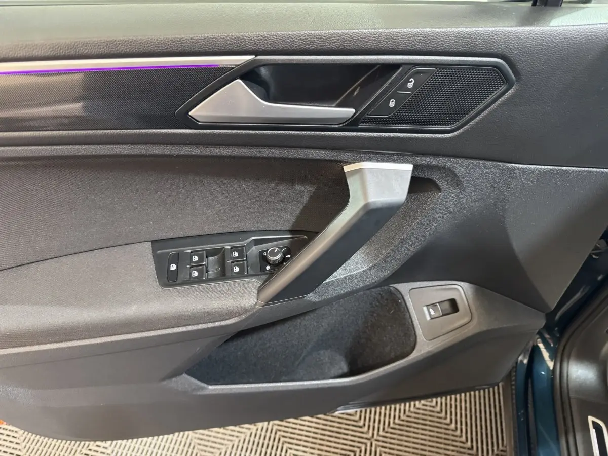 Vue rapprochée de la porte avant gauche du Volkswagen Tiguan bleu, montrant les commandes des vitres et la poignée intérieure.