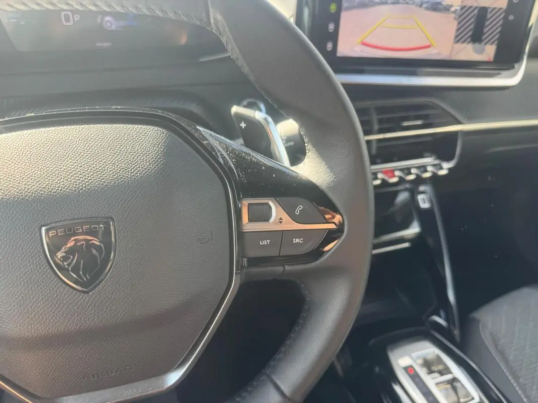 Gros plan sur le volant cuir noir de la Peugeot 2008 Hybrid 145, avec commandes intégrées et écran tactile en arrière-plan.