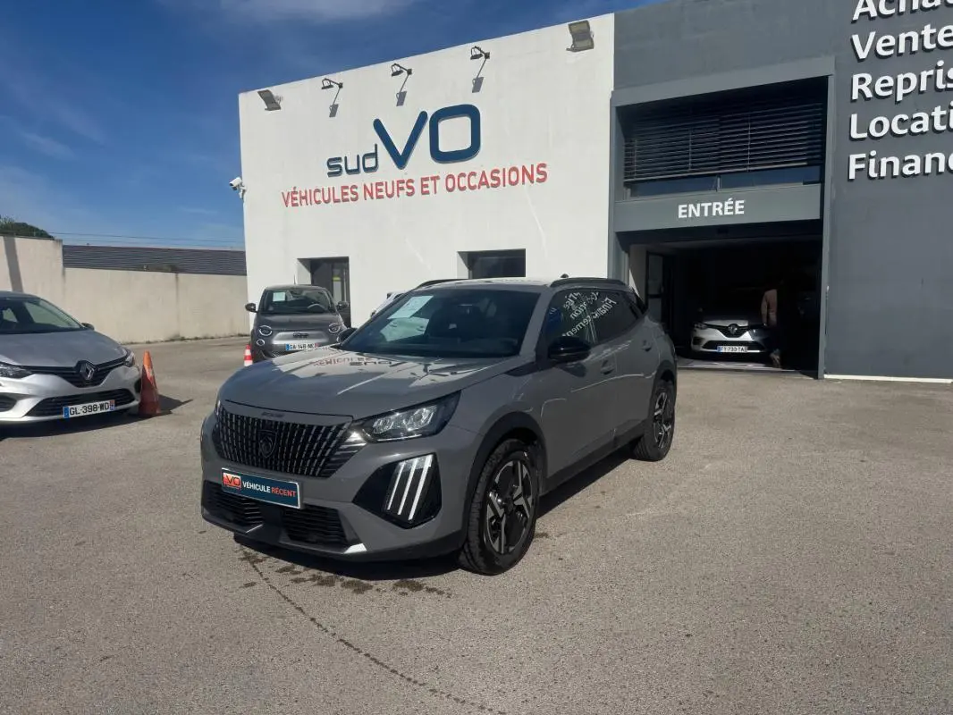 Peugeot 2008 Hybrid 145 gris vue 3/4 avant droit, jantes alliage 17 pouces, calandre noire distinctive, stationné devant un garage.