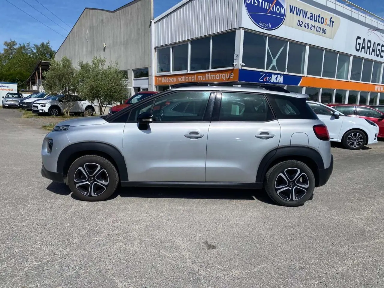 Vue de profil côté gauche d'un Citroën C3 Aircross gris Artense avec toit noir et jantes distinctives, stationné devant un garage.