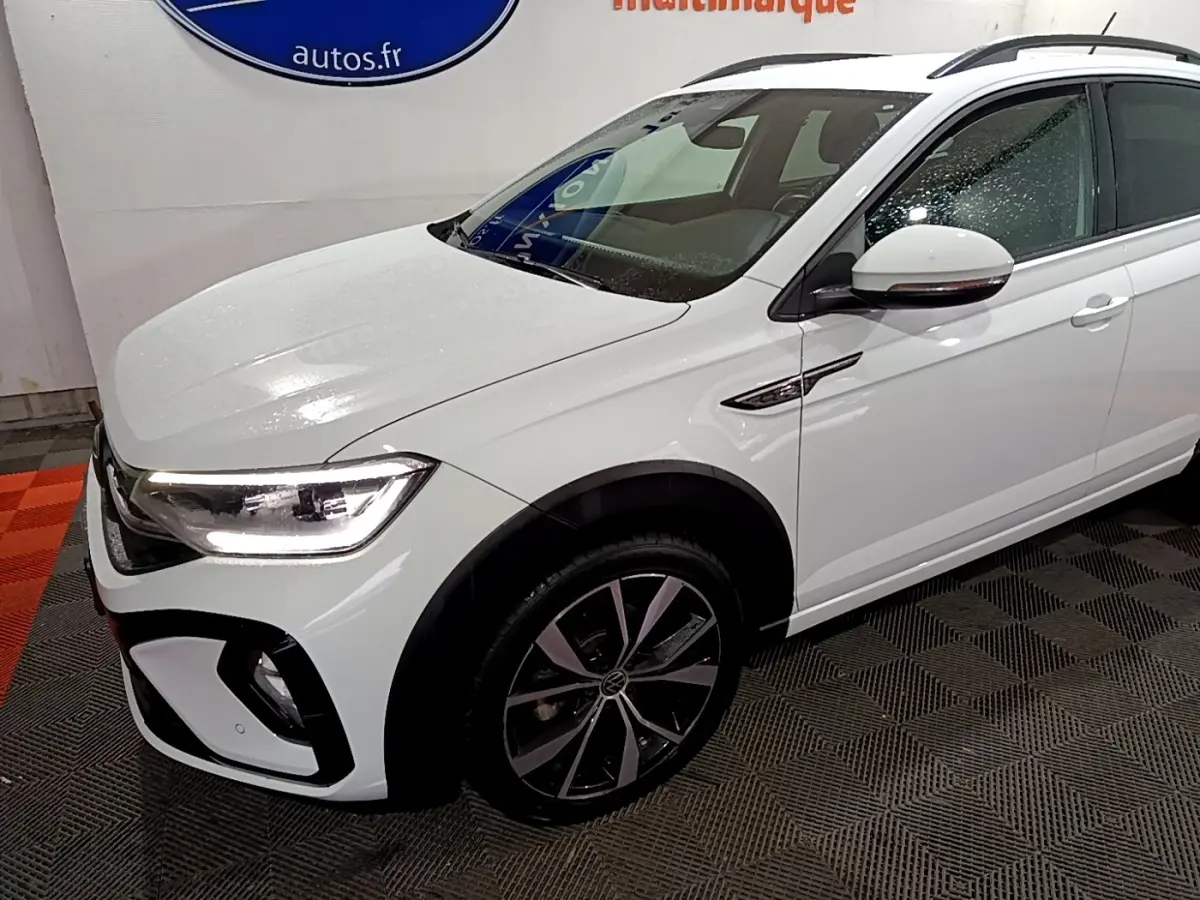 Vue 3/4 avant droite du Volkswagen Taigo blanc 1.0 TSI 110 DSG R-Line avec jantes alliage et feux LED allumés.