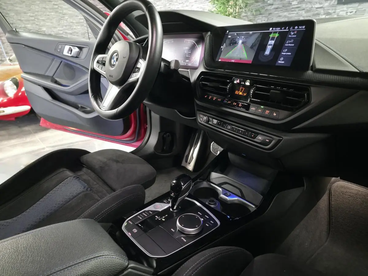 Vue intérieure côté conducteur d'une BMW Série 1 116d rouge, avec console centrale moderne et écran tactile affichant la caméra de recul.