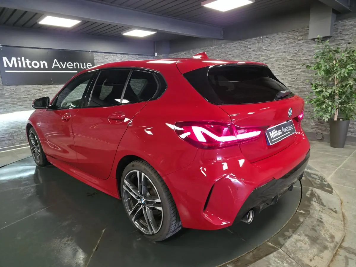 BMW Série 1 116d M Sport rouge vue 3/4 arrière droit, avec jantes alliage 18 pouces et feux LED allumés.