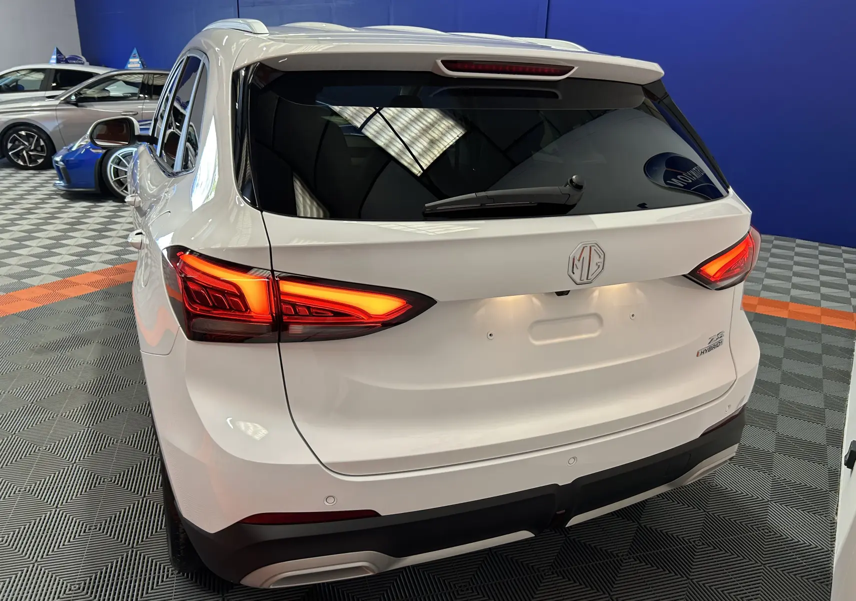 Vue 3/4 arrière droite du MG ZS 1.5 Hybrid+ 197 Luxury en blanc Dover avec feux arrière allumés dans un showroom.