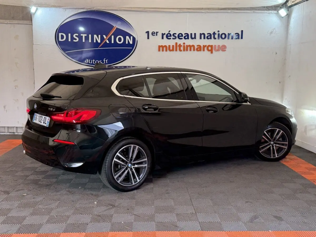 BMW Série 1 116d noir vue 3/4 arrière droit dans un showroom avec logo Distinxion en arrière-plan.