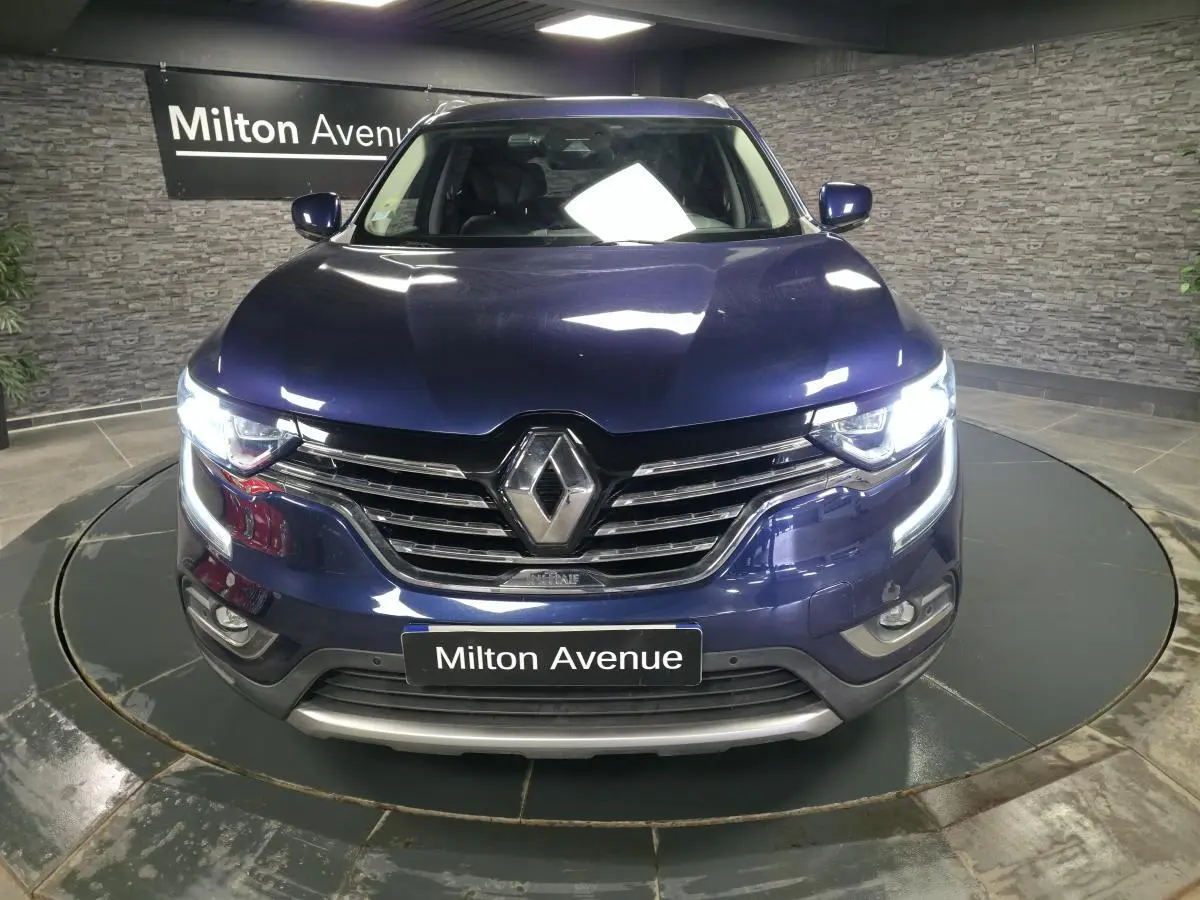 Vue frontale d'un Renault Koleos bleu avec calandre chromée et feux LED allumés en intérieur showroom.