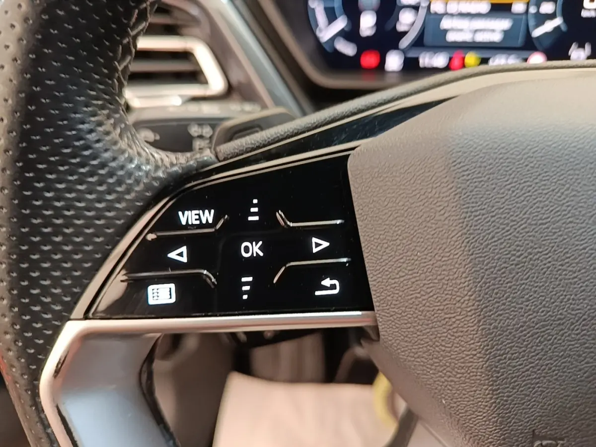 Gros plan sur les commandes gauche du volant cuir perforé de l'Audi Q4 E-TRON Sportback noir, avec affichage numérique flou en arrière-plan.