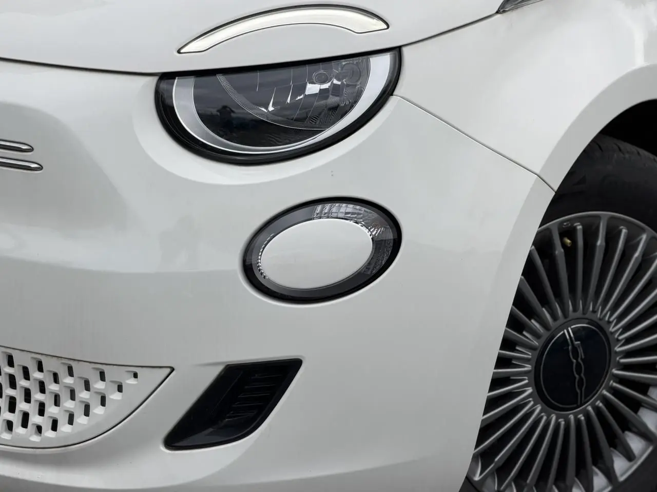 Gros plan sur l'avant droit blanc d'une FIAT 500 électrique 2023, mettant en valeur le phare LED et la jante stylisée.