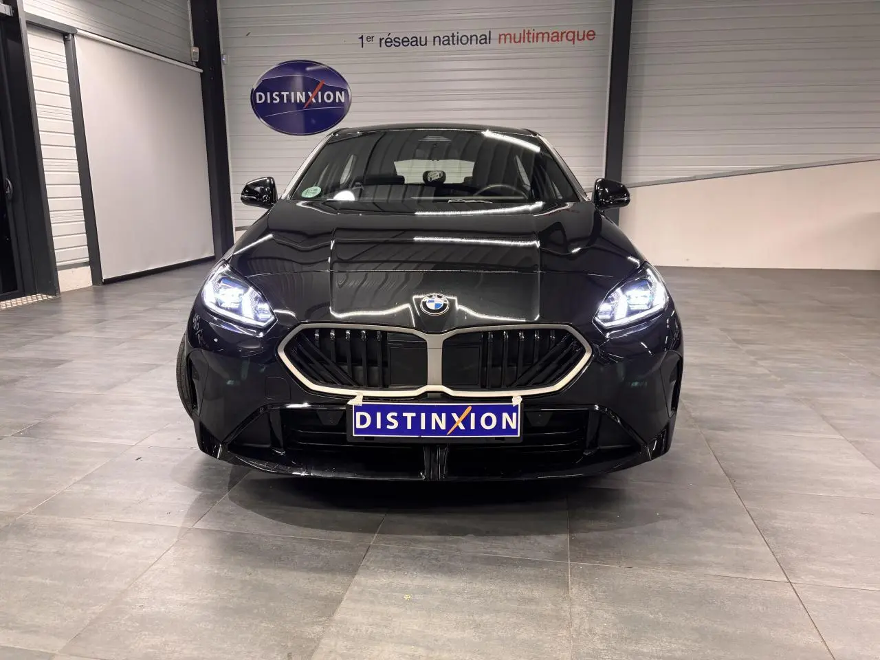 BMW Série 1 118d M Sport noir métal vue de face avec calandre chromée et phares LED allumés dans un showroom.