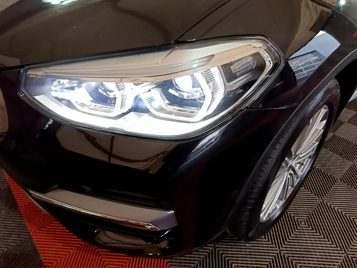 Gros plan sur le phare avant droit allumé d’un BMW X3 noir, avec jante alliage visible et carrosserie brillante.