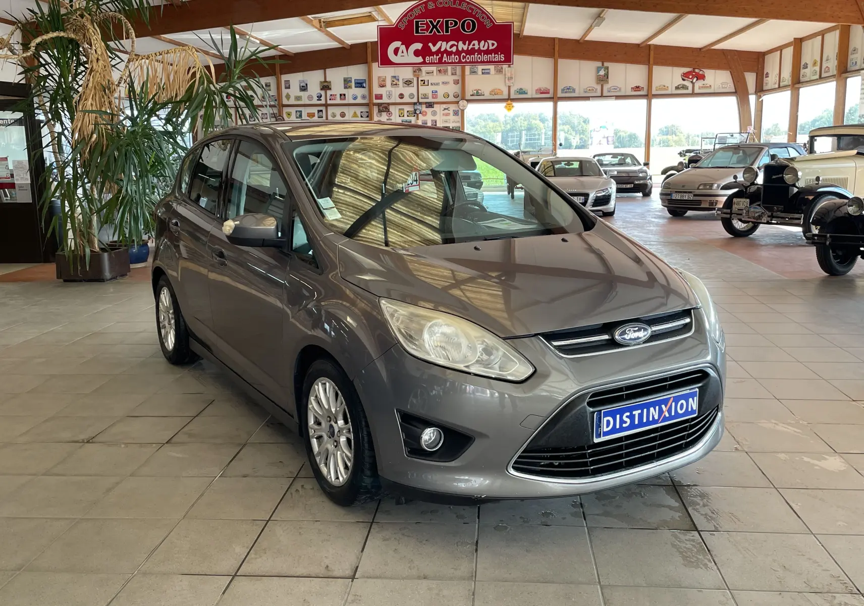 Ford C-MAX gris vue 3/4 avant droit dans un showroom avec plaque Distinxion visible sur le pare-chocs.