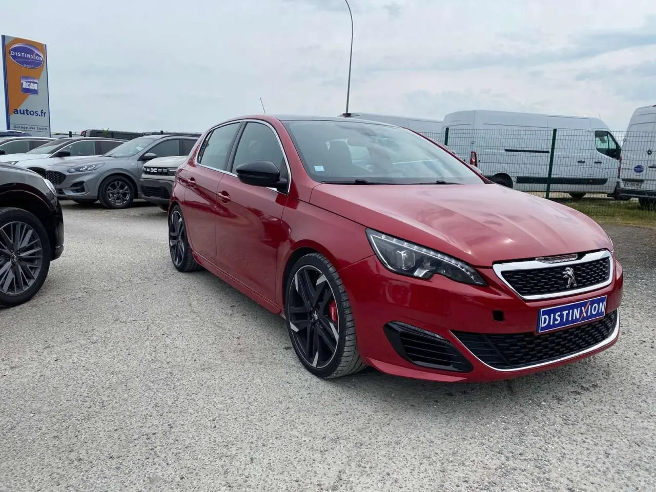Peugeot 308 GTi rouge vue 3/4 avant avec jantes noires et calandre chromée sur parking extérieur.