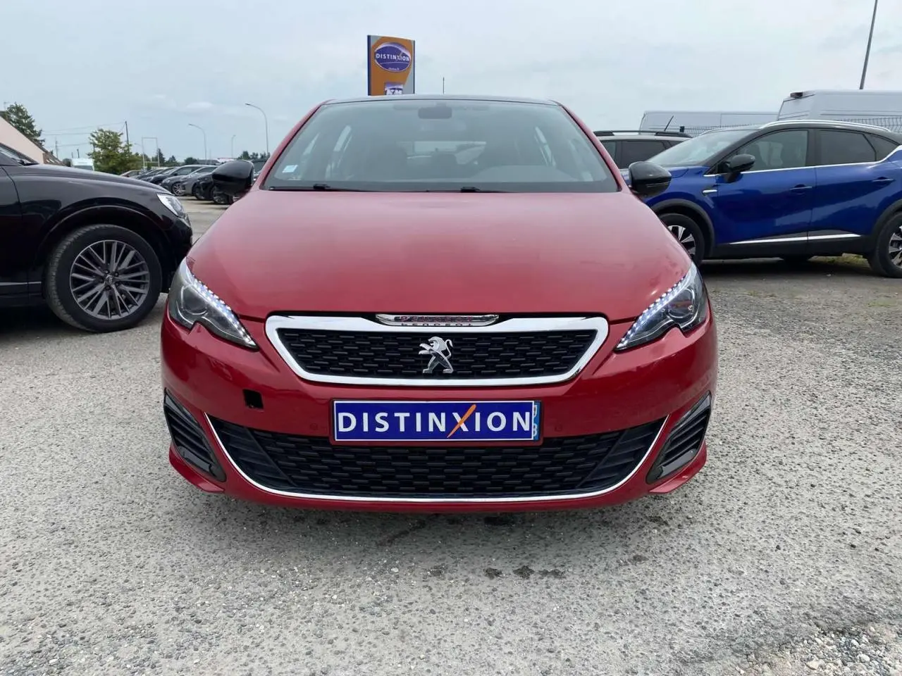 Vue frontale d'une Peugeot 308 GTi rouge avec calandre chromée et phares LED allumés sur parking.
