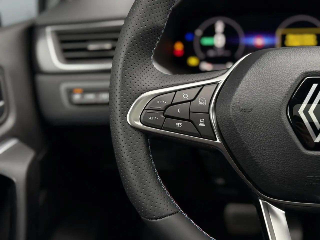 Gros plan sur le volant perforé avec commandes intégrées et logo Renault, tableau de bord numérique flou en arrière-plan.