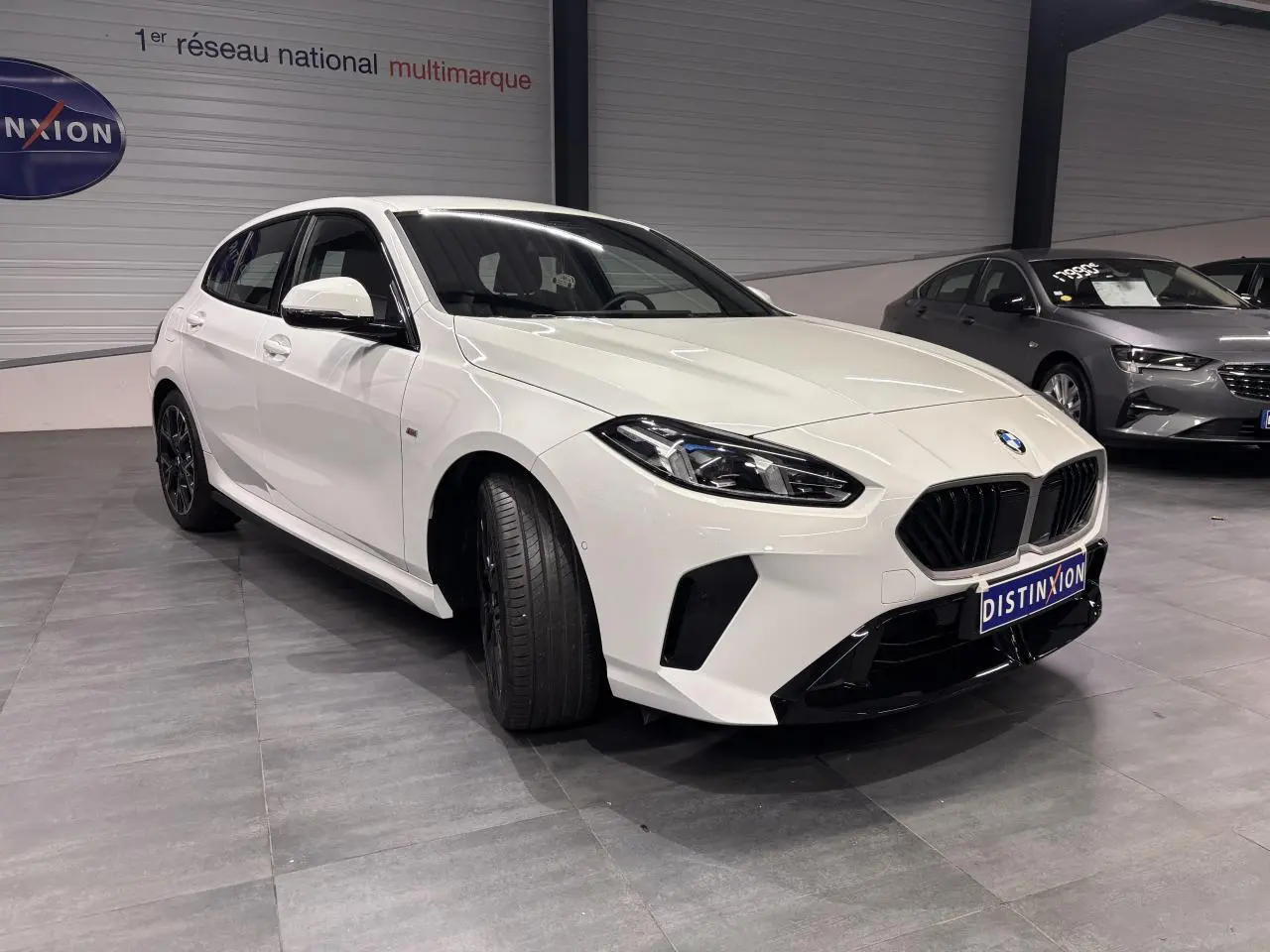 BMW Série 1 118D M Sport blanche vue 3/4 avant gauche avec calandre noire et jantes 18 pouces bicolores.