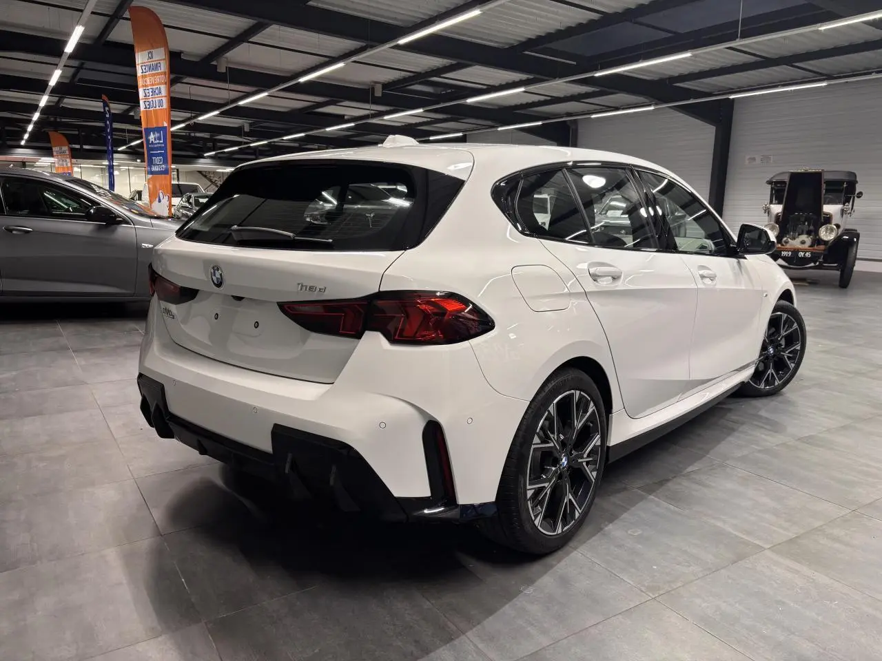 BMW Série 1 118D blanche vue 3/4 arrière droit en intérieur, avec jantes 18 pouces et éléments M Sport noirs.