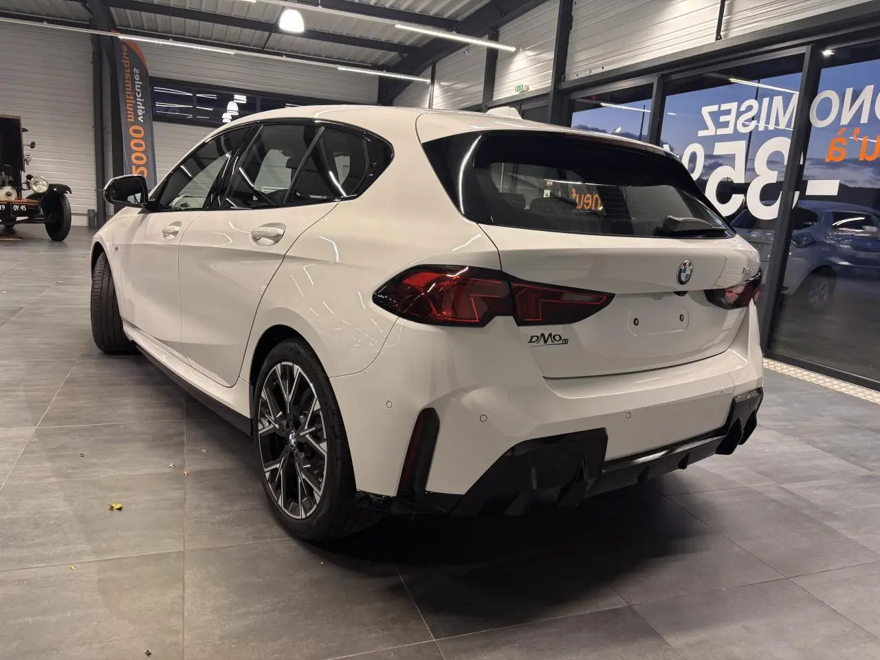 Vue 3/4 arrière droite d'une BMW Série 1 118D blanche en finition M Sport avec jantes 18 pouces en alliage bicolore.