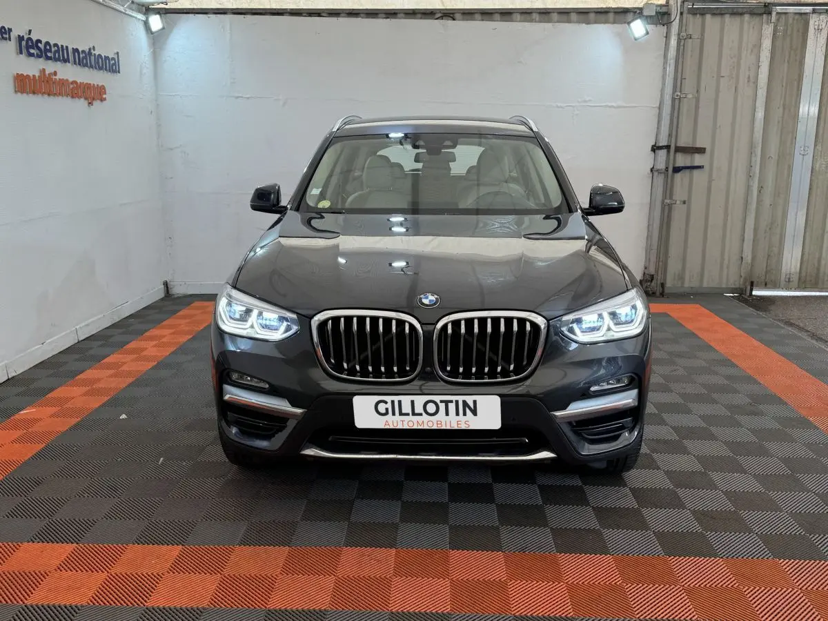 BMW X3 xDrive30d gris foncé vue de face avec phares LED allumés dans un showroom au sol noir et orange.
