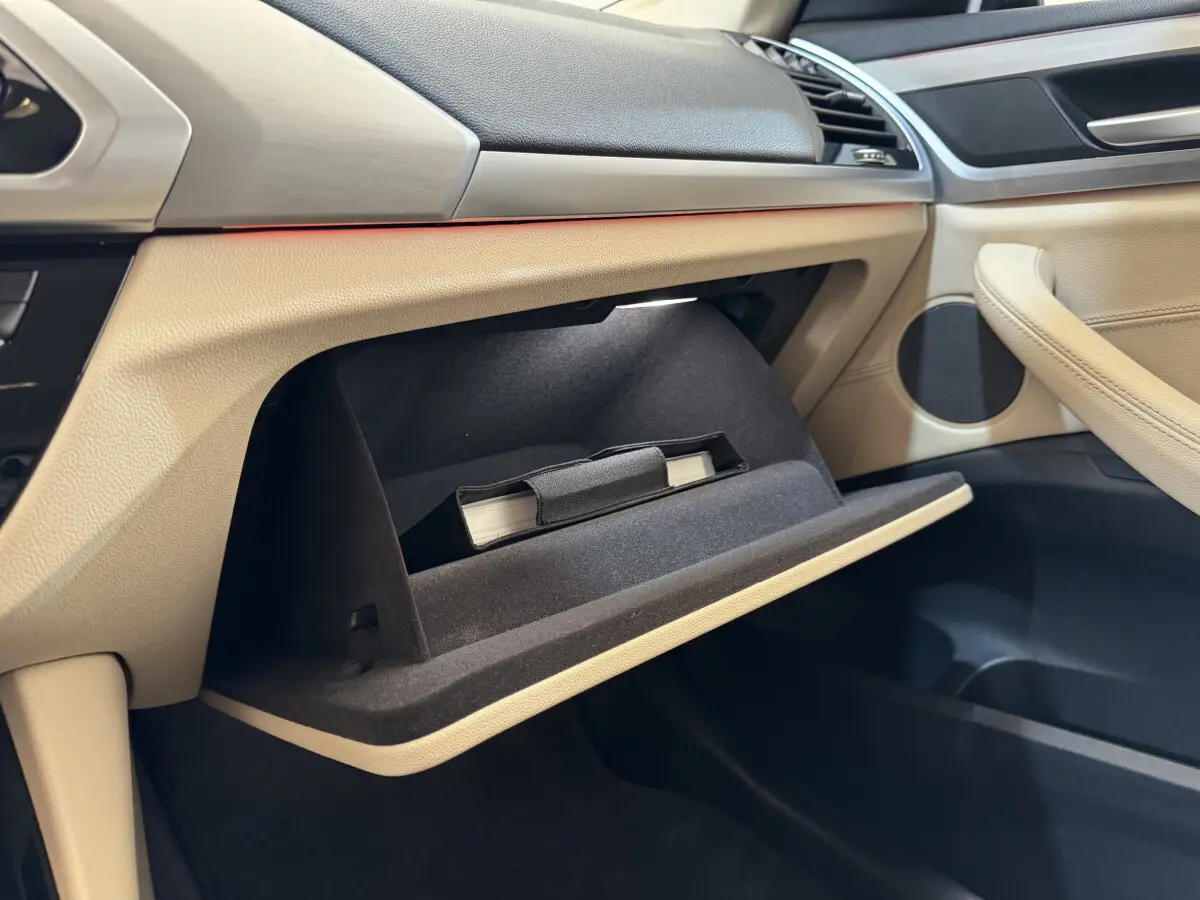 Intérieur beige clair du BMW X3 2018, vue côté passager sur la boîte à gants ouverte avec manuel visible.