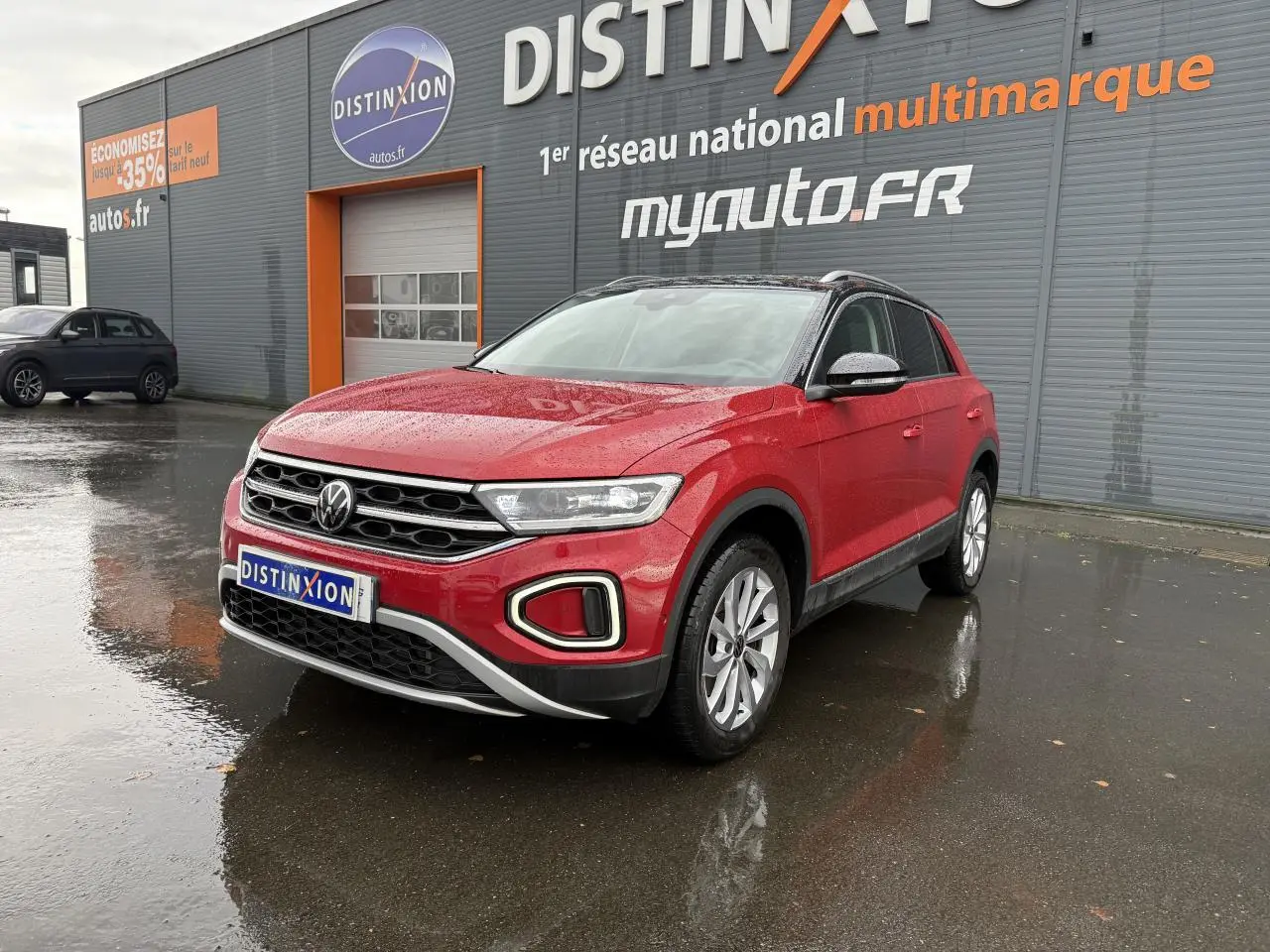 Volkswagen T-Roc rouge Kings Red métallisé en 3/4 avant droit, garé devant un bâtiment gris avec enseigne Distinxion.