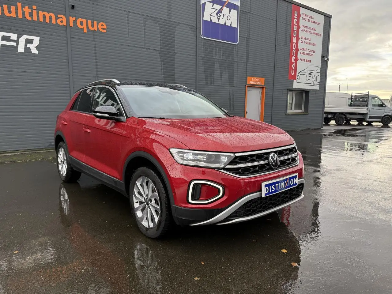 Volkswagen T-Roc rouge Kings Red métallisé vu en 3/4 avant droit sur sol mouillé devant un bâtiment gris.