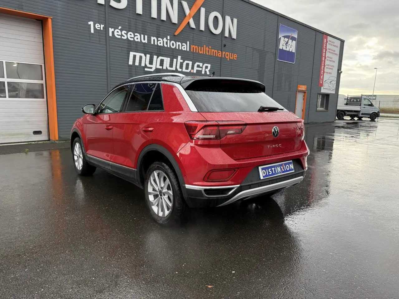 Volkswagen T-Roc rouge Kings Red métallisé vu en 3/4 arrière droit sur sol humide devant un bâtiment gris.