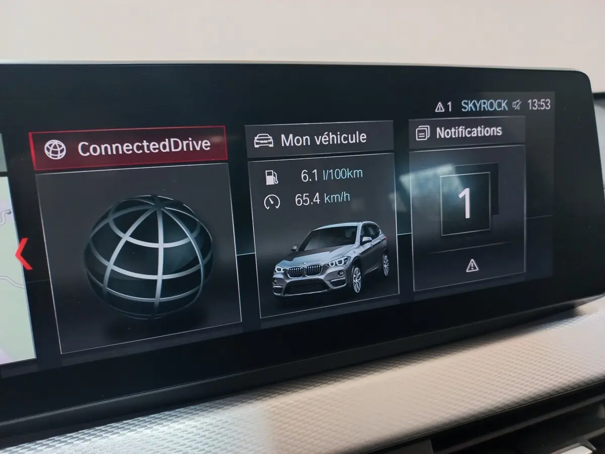 Écran tactile intérieur montrant la consommation et la vitesse d'un BMW X1 noir en vue 3/4 avant.