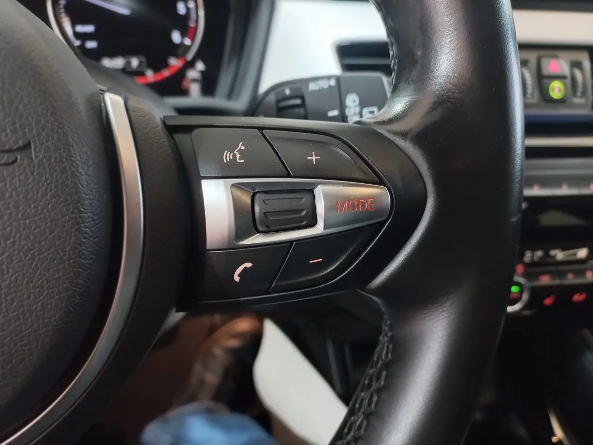 Gros plan sur les commandes du volant cuir noir de la BMW X1 xDrive20d M Sport, avec bouton mode rouge visible.
