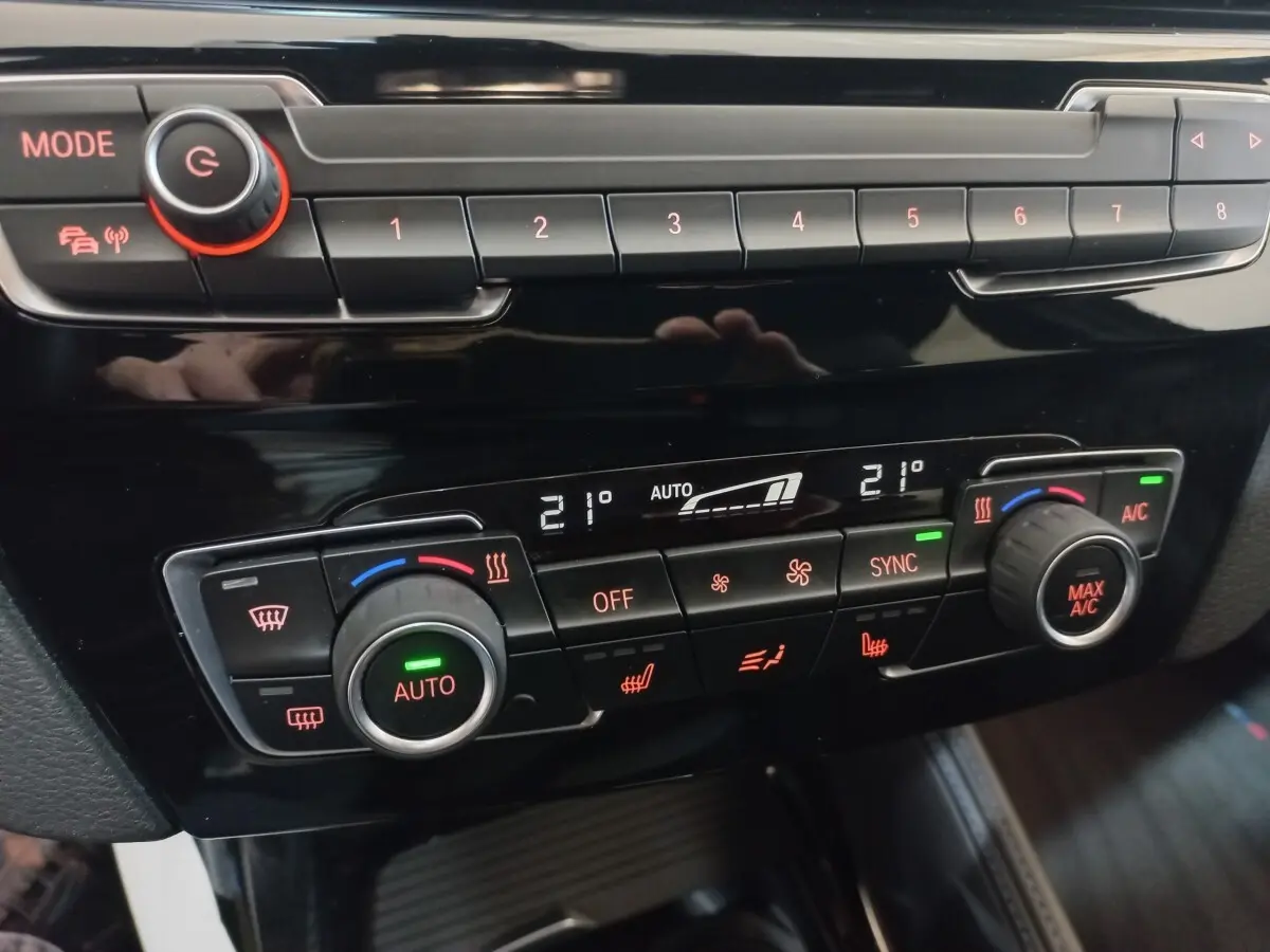 Gros plan sur la console de climatisation digitale de la BMW X1 noire, avec réglages à 21°C et boutons rétroéclairés rouges.