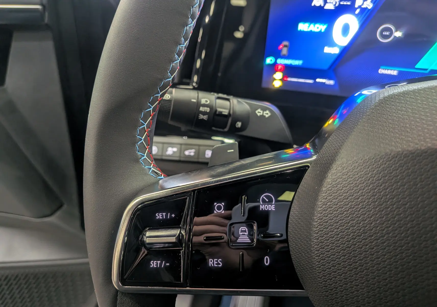 Gros plan sur le volant cuir noir avec surpiqûres tricolores et commandes intégrées, tableau de bord numérique en arrière-plan.