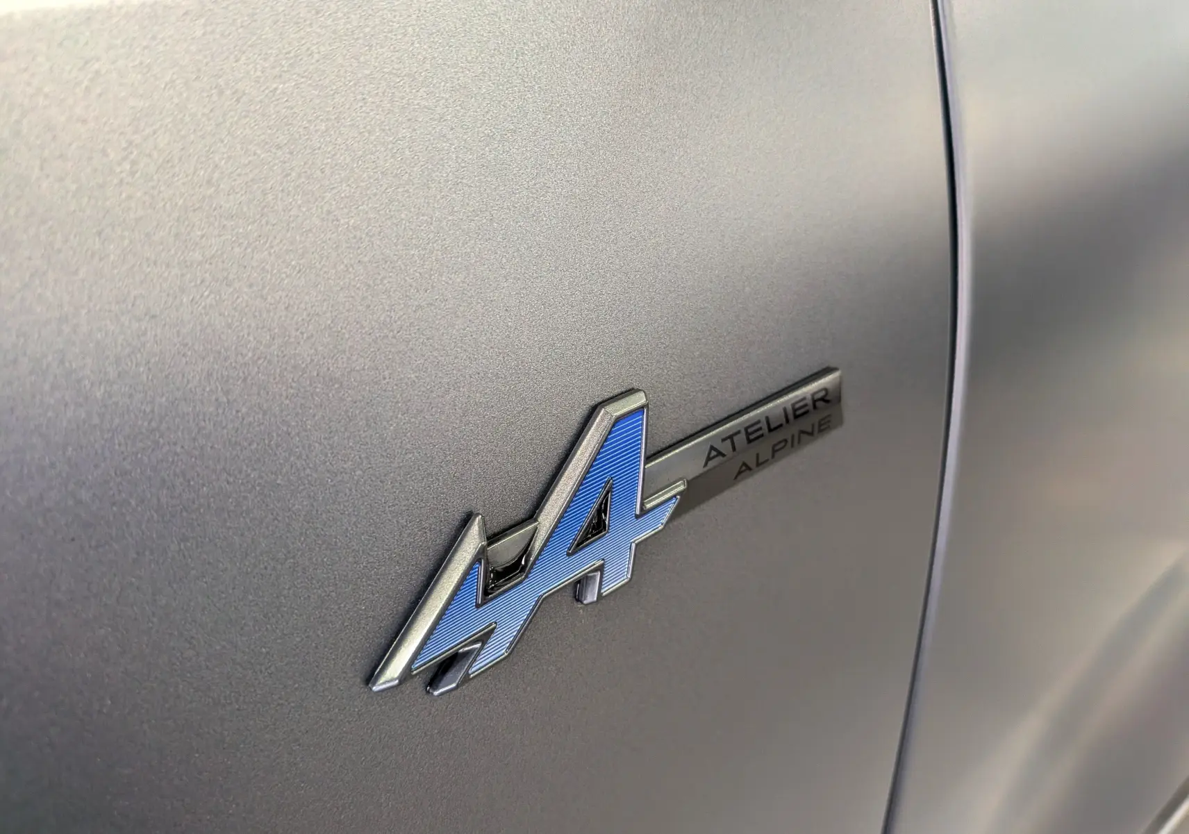 Gros plan sur l’insigne bleu et argent "4 Atelier Alpine" sur la carrosserie gris schiste satin du Renault Rafale.