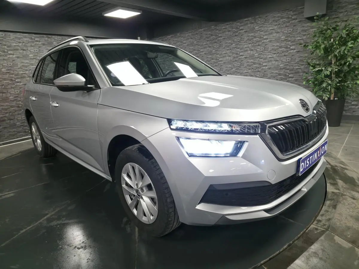 Vue 3/4 avant droit d'un Skoda Kamiq gris métallisé avec feux LED allumés dans un showroom intérieur.