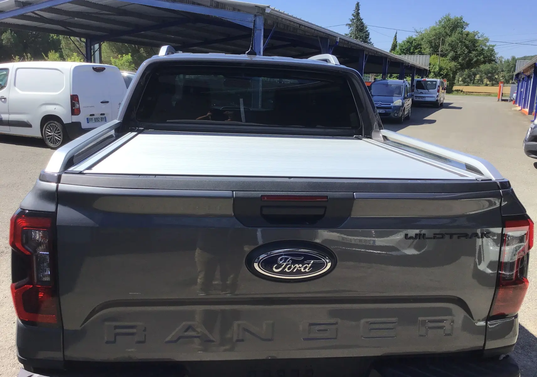 Vue arrière d'un Ford Ranger Double Cabine gris carbone avec rideau de plateau coulissante électrique et logo Wildtrak visible.