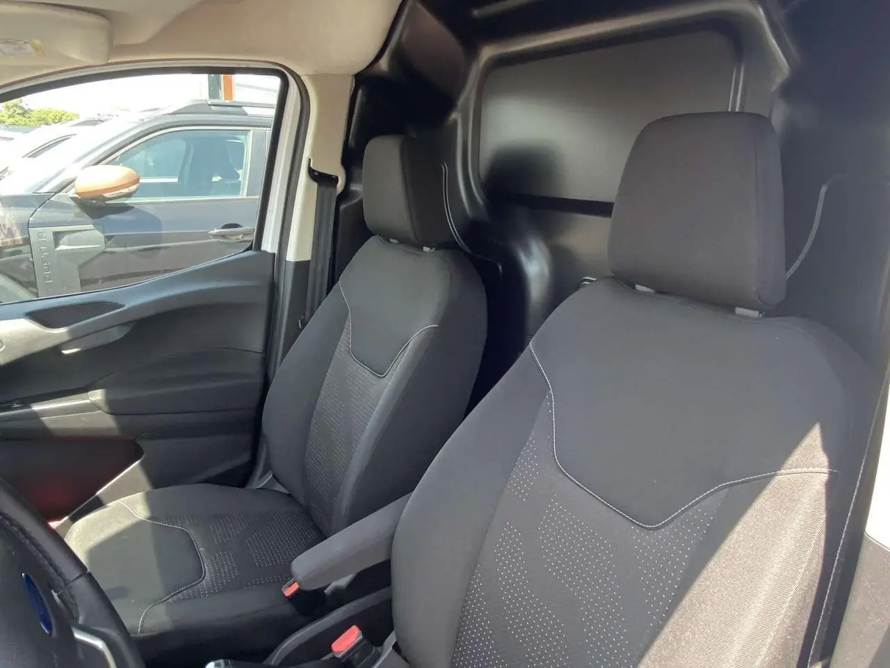 Vue intérieure côté conducteur et passager du Ford Tourneo Courier blanc, montrant les sièges noirs et la cloison tôlée arrière.