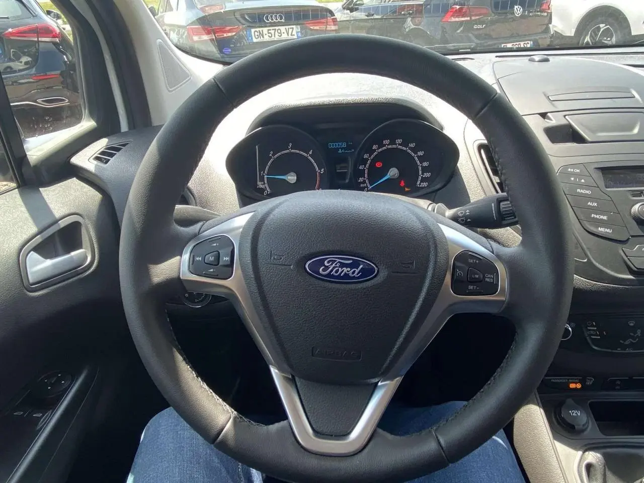 Vue intérieure centrée sur le volant cuir Ford d'un Tourneo Courier blanc, avec tableau de bord et commandes au volant visibles.