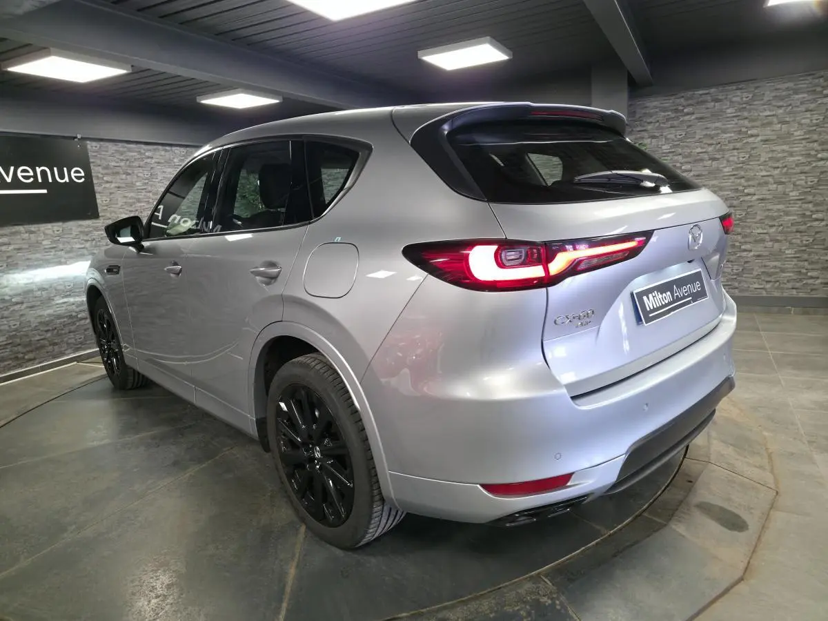 Vue 3/4 arrière droite du Mazda CX-60 gris Sonic Silver avec jantes noires et feux arrière LED allumés en intérieur showroom.