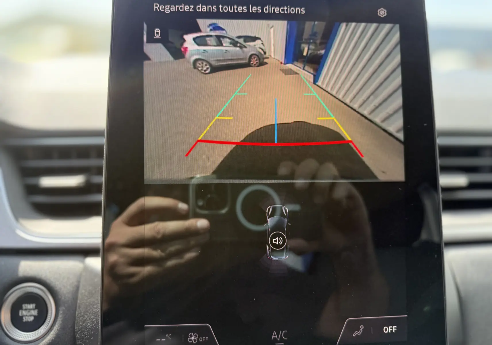 Vue caméra de recul montrant une voiture grise stationnée côté droit devant un bâtiment avec marquages au sol colorés.