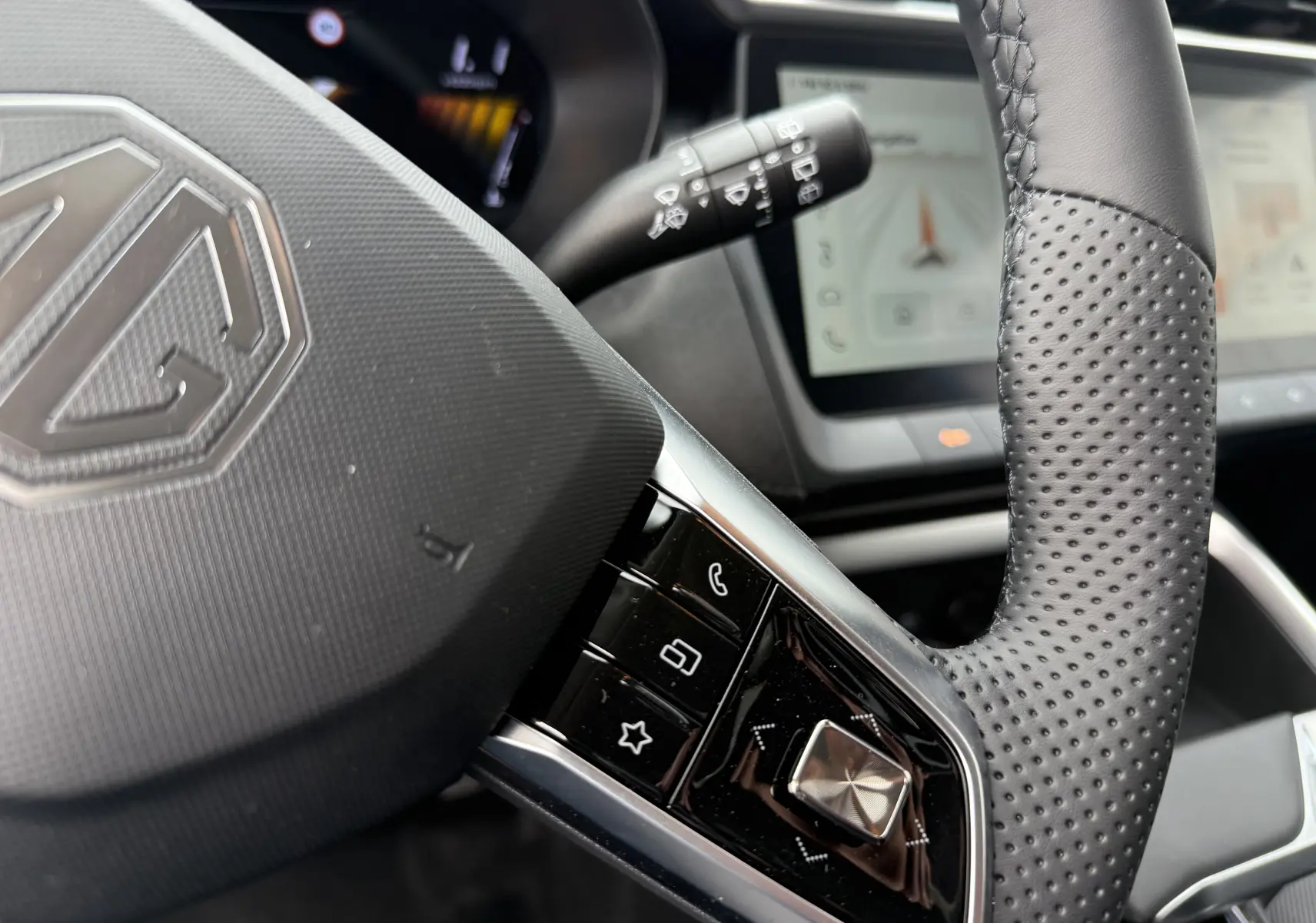 Gros plan sur le volant perforé noir du MG MOTOR ZS 1.5 HYBRID+ 197 LUXURY avec commandes intégrées et tableau de bord numérique flou.