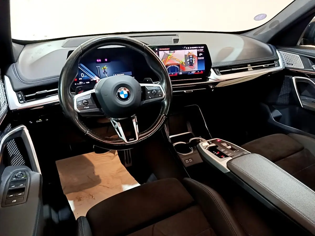 Intérieur BMW X1 xDrive23i M Sport 2023 vu côté conducteur, tableau de bord noir avec écran tactile et volant cuir alu.