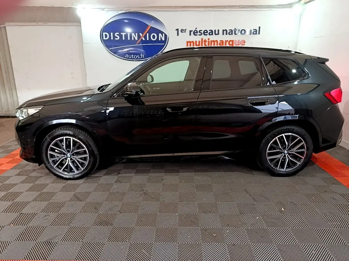 BMW X1 xDrive23i M Sport noir vue de profil côté gauche, jantes alliage et logo Distinxion en arrière-plan.