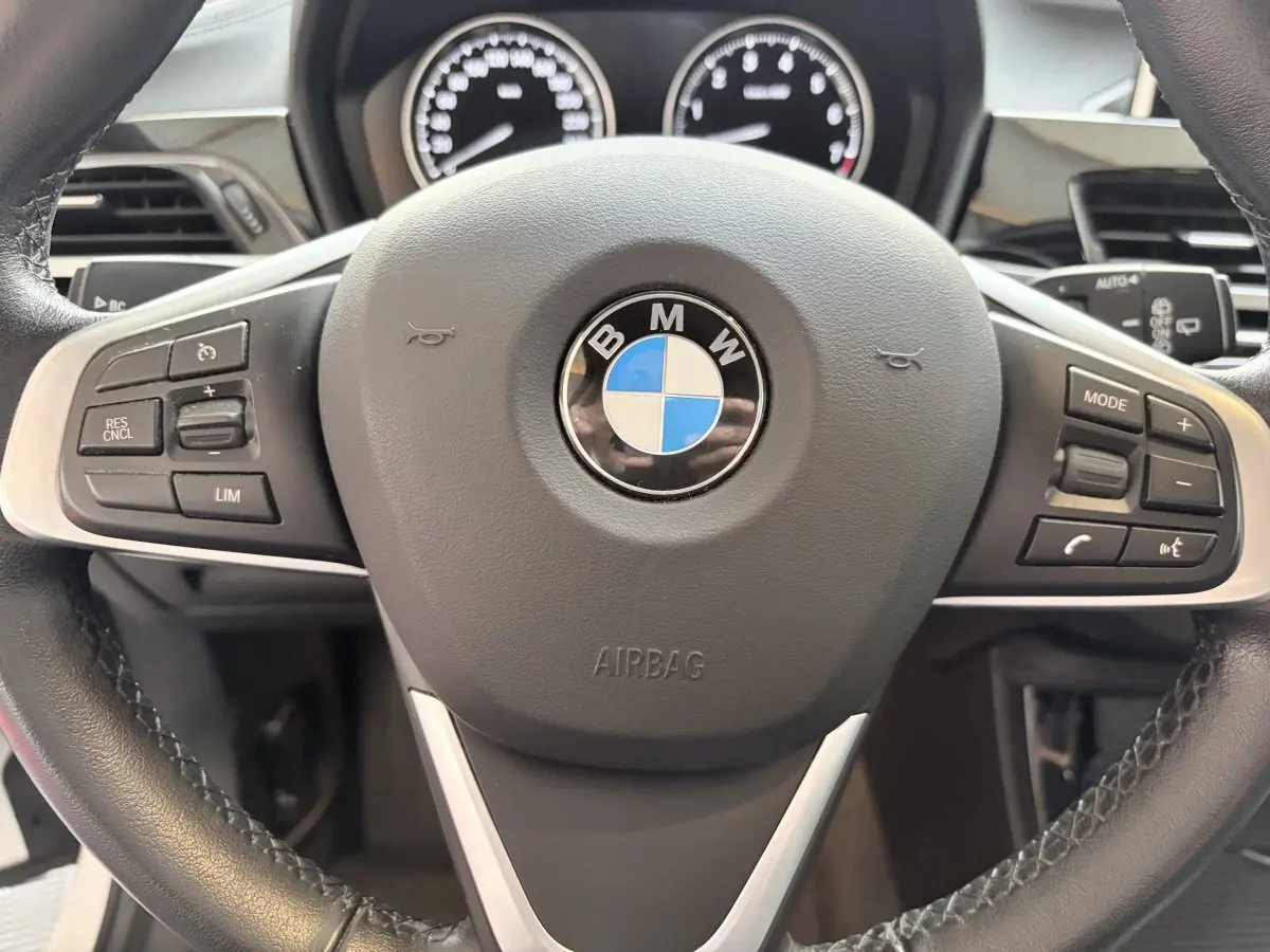 Vue rapprochée du volant cuir et alu multifonctions d'une BMW X1 blanche avec tableau de bord numérique en arrière-plan.