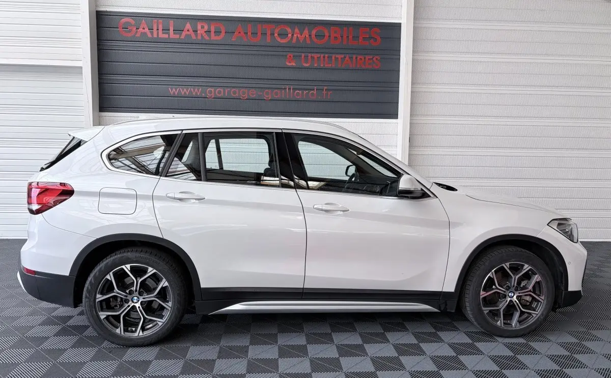 Profil droit d’un BMW X1 blanc 2021 avec jantes alliage et rails de toit chromés, stationné en intérieur.