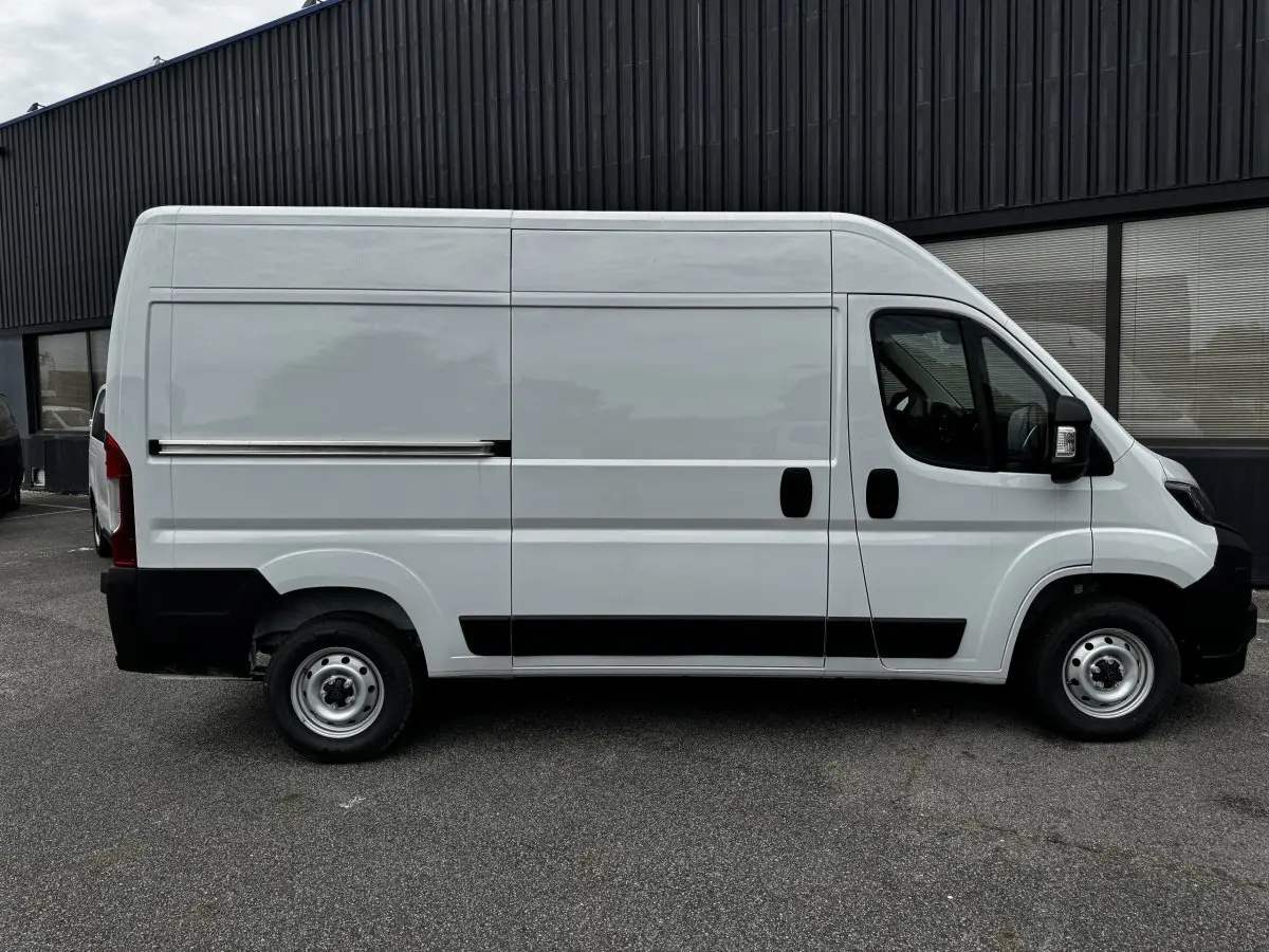 Vue de profil côté droit d'un fourgon Peugeot Boxer blanc avec porte coulissante et protections noires sur un sol goudronné.